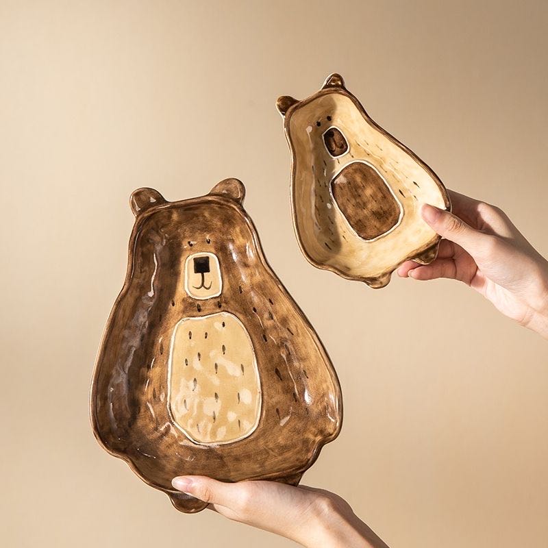 Two bear-shaped ceramic plates held by hands against a beige background