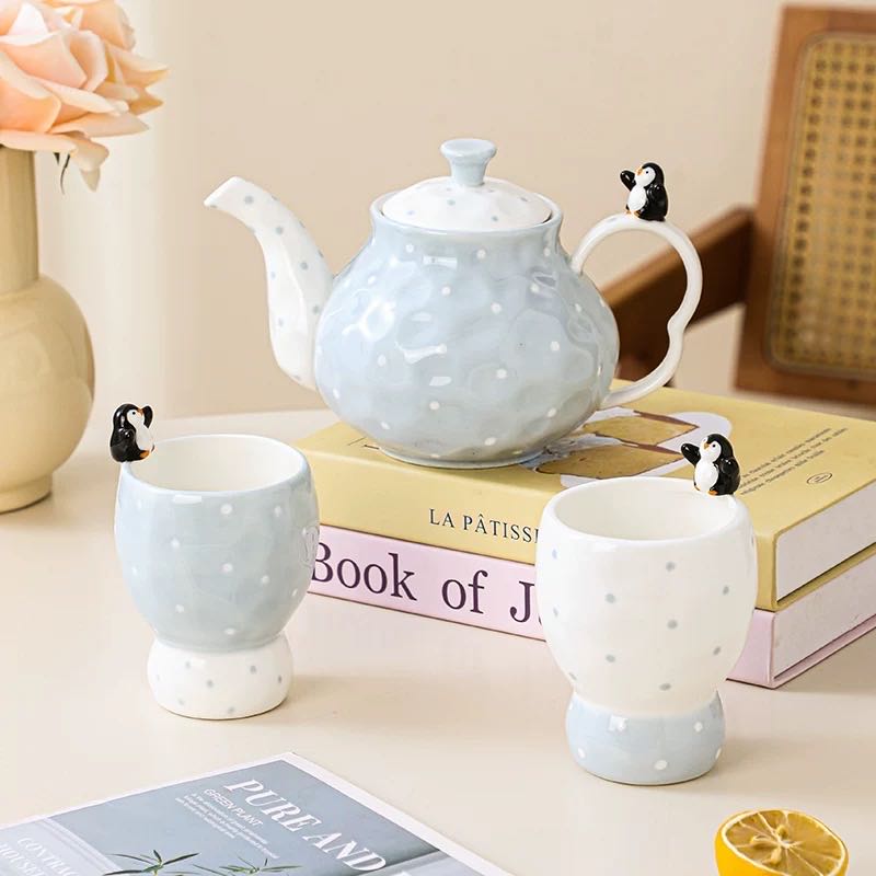 Ceramic teapot and mugs with penguin handles on a table with books and a vase.
