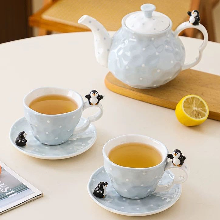Ceramic teapot and cups with penguin handles on a table with lemon and magazine.