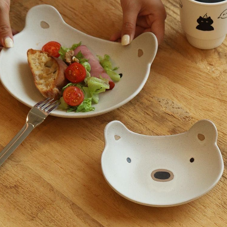 Person holding a plate with salad and bread, next to a cat-shaped ceramic dish on a wooden table.