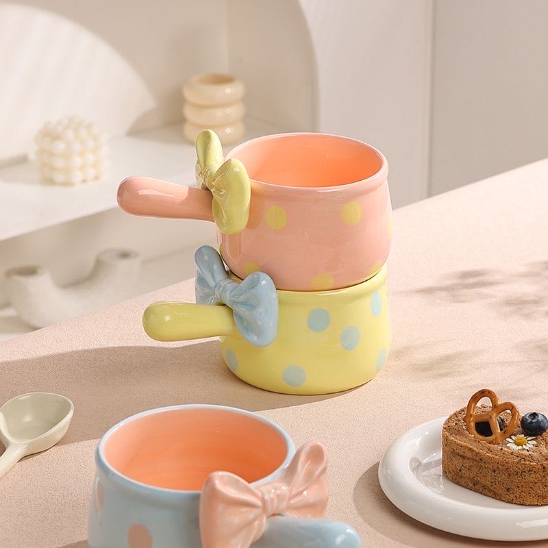 Set of colorful ceramic mugs with bow designs on a table with a dessert.