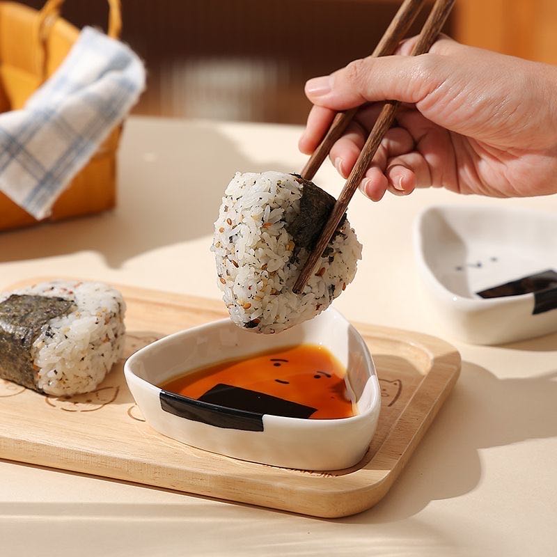 Person dipping a sushi ball into soy sauce using chopsticks on a wooden board.