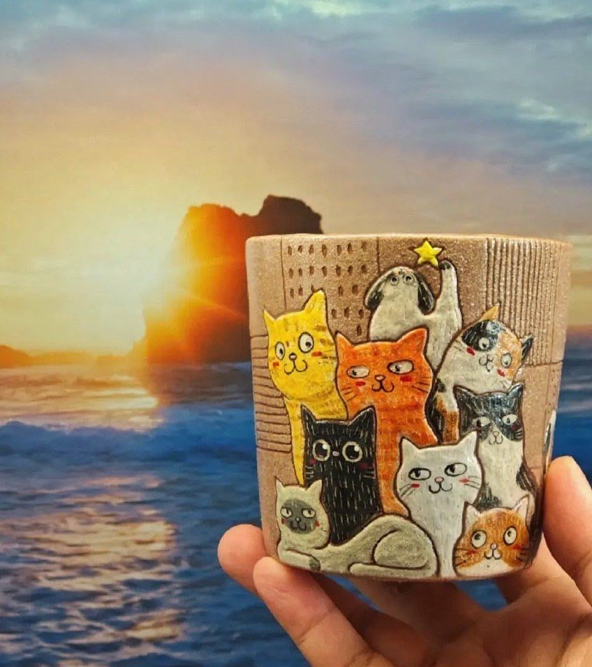 Cup with cat illustrations held in front of a scenic sunset over water