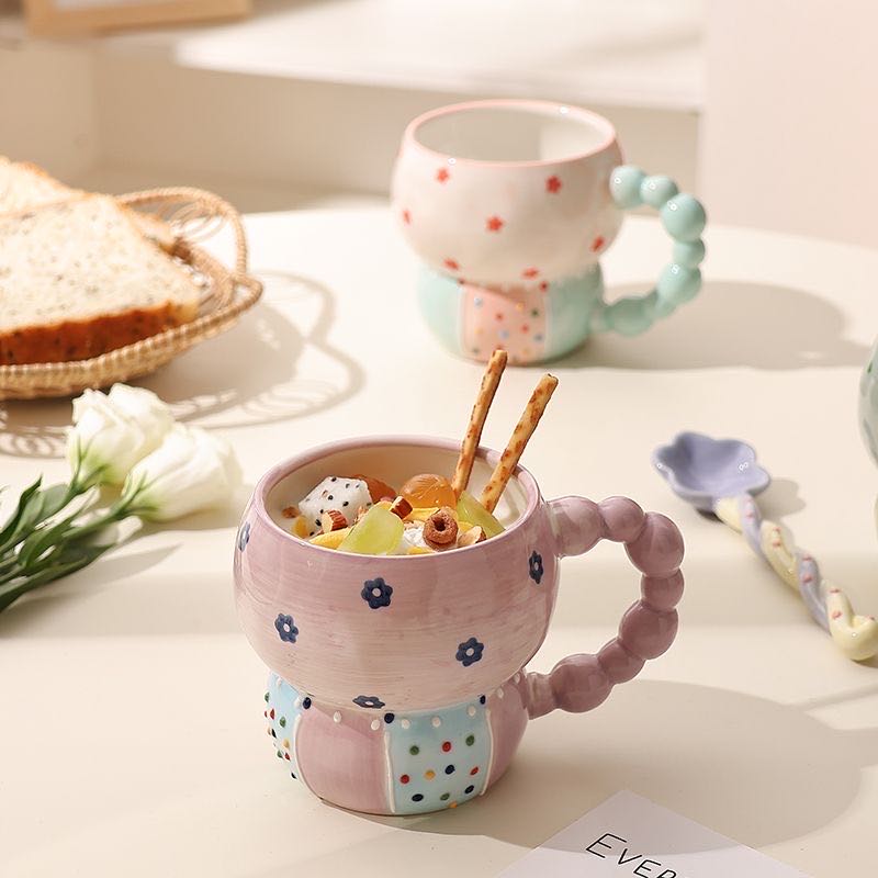 Decorative mug with floral patterns filled with cereal and fruit, placed on a table with bread and flowers.