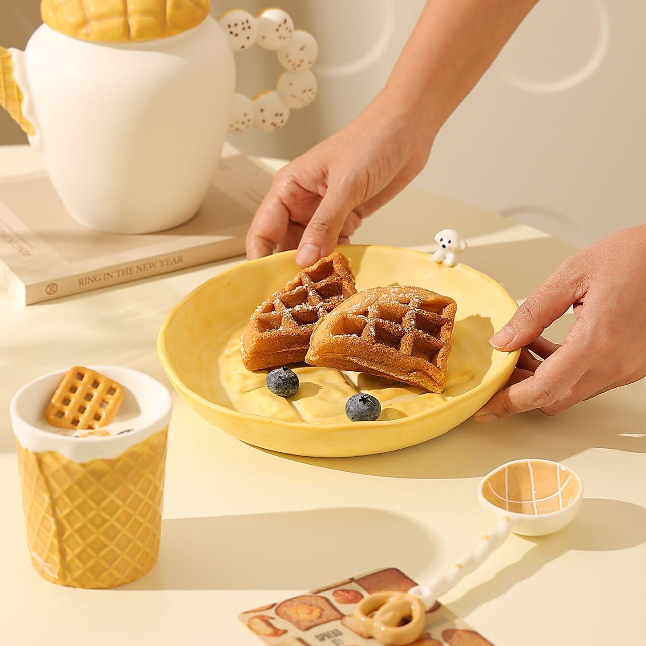 Person arranging waffles on a yellow plate with a decorative teapot and small dog figurine in the background.